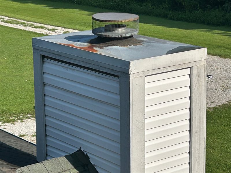 A sided chimney chase seen from the roof. Its flat metal cap is streaked with rust around the flue, and the cap's edges show staining where water has been running off.