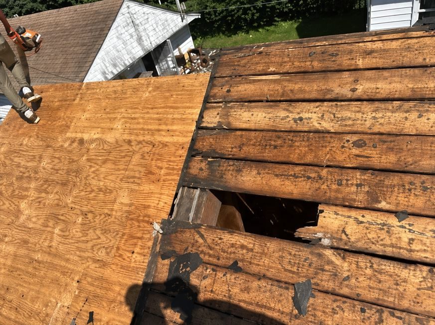 Roof decking exposed after tear-off: rotted plank sheathing with a large hole punched through it into the attic on the right, and fresh plywood already laid on the left.