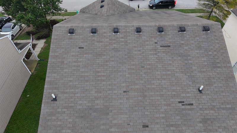 Aerial view straight down a large asphalt shingle slope. A row of seven box vents runs along the ridge, two pipe boots stand in the field, and roughly a dozen individual tabs scattered across the slope are darker than the shingles around them.