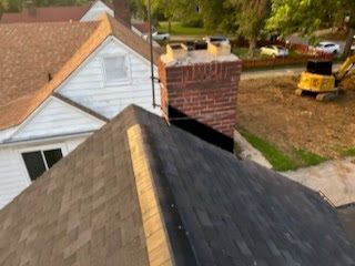 The finished roof from the ridge: new architectural shingles and ridge caps running past a brick chimney, with the neighborhood behind.