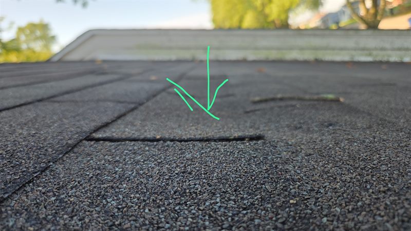 A green inspector's arrow pointing at a shingle tab whose edge has lifted off the course below it.