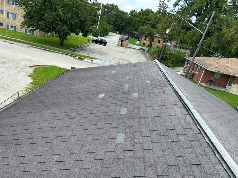 Looking down a dark asphalt shingle slope toward a parking lot. About a dozen individual tabs across the field are a lighter gray than the shingles around them, each one a replacement shingle.